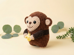 A crocheted plush monkey toy holding a yellow banana, standing against a warm cream background with decorative eucalyptus leaves.
