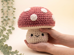 A crocheted mushroom plush toy with a dusty pink cap, white polka dots, and a cream-colored stem with a cute embroidered face, being held in two hands against a warm beige background with a decorative eucalyptus branch.