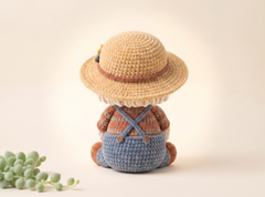 A crocheted doll viewed from the back, wearing a straw-style hat with a brown band, a peach and blue striped shirt, and blue overalls, set against a warm cream background with a small green plant in the lower left corner.