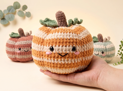 A hand holds a crocheted pumpkin-shaped plush toy in the foreground, with two more similar toys blurred in the background against a soft cream backdrop with decorative leaves.