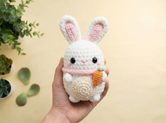 A hand holds a white crocheted bunny plush toy with pink inner ears, black bead eyes, a pink nose and scarf, and an orange carrot accessory, against a warm beige background with blurred green plants.