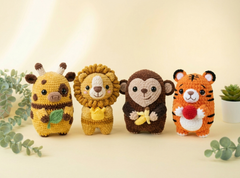 Four crocheted animal plush toys arranged in a row on a light beige surface with soft greenery props. The animals are a yellow cow holding a green leaf, a yellow lion with a tan mane, a brown monkey holding a yellow banana, and an orange tiger holding a red apple.