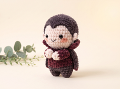 A small crocheted doll with dark brown hair, a cream face and hands, and a dark burgundy dress, standing against a warm cream background with a sprig of green leaves on the left.
