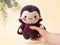 A hand holding a small crocheted doll with a dark purple hair and dark burgundy outfit against a warm beige background with a leafy plant in the corner.