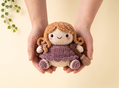 A crocheted doll with brown hair and a purple dress is held in two hands against a soft beige background with a small green plant in the corner.