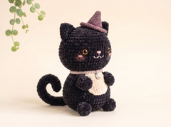 A crocheted black cat plush toy wearing a small pointed witch hat and a cream bib, posed against a warm beige background with a decorative green leafy vine in the upper left corner.