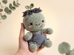 A handmade crocheted doll with a green head, dark gray yarn hair, and blue-gray overalls, held in a person's hand against a soft beige background with blurred green leaves.
