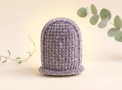 A small crocheted dome-shaped item in a soft lavender-gray color, photographed on a warm cream background with decorative green leaves in the corners.