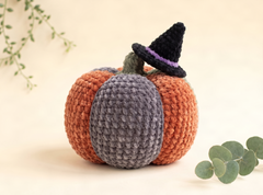A crocheted pumpkin decoration with orange sides, a gray center panel, a dark green stem, and a black witch hat with a purple band, styled on a warm cream background with eucalyptus leaves.