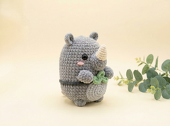 A crocheted gray rhinoceros toy with a small beige horn, pink nose, and green leaf detail, standing on a warm beige background with eucalyptus branches to the right.