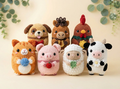 Six crocheted animal plush toys arranged together on a light beige surface with a soft decorative background and eucalyptus leaves. The toys include an orange cat holding blue yarn, a pink pig holding a red strawberry, a white sheep holding a green clover, and a black-and-white cow.