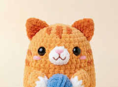 A close-up of a crocheted orange tabby cat toy with black bead eyes, a white muzzle, pink cheeks, and a blue ball of yarn in its paws, set against a plain beige background.