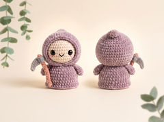 Two crocheted amigurumi doll figures in a lavender-purple hooded outfit, one shown from the front holding a small gray scythe and the other from the back, on a warm cream background with soft green eucalyptus leaves.