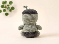 A handmade crocheted amigurumi doll shown from the back, featuring a light sage green body with dark gray stripes on the hat and bottom, set against a warm cream background with a small green plant in the corner.