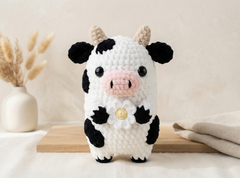 A crocheted cow plush toy with black and white patches, a pink snout, beige horns, and a small white flower with a gold center on its chest, standing on a wooden board in a softly styled indoor setting.