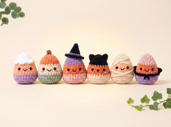 Six cute crocheted pumpkin-shaped characters lined up on a warm cream background with decorative green leaves in the corners. Each character has a different Halloween-themed color scheme and facial expression.