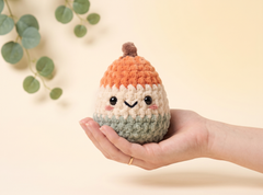 A small crocheted amigurumi character doll with a smiling face, held in a person's hand against a soft beige background with blurred green leaves in the corner.