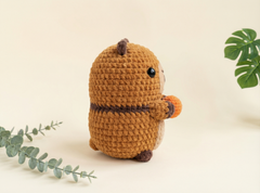 A crocheted capybara plush toy in warm brown yarn, holding an orange carrot, styled with eucalyptus and monstera leaves on a light beige background.