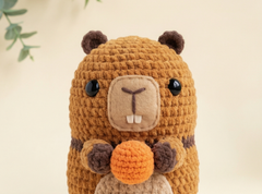 A crocheted beaver plush toy holding an orange nut, set against a soft beige background with subtle green leaves in the corner.