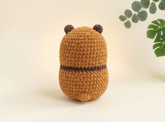 A crocheted bear-shaped plush toy in warm brown with darker brown ear accents and a horizontal stripe, photographed on a light beige background with green leaves in the upper right corner.