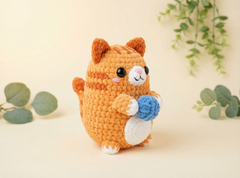 A crocheted orange cat plush toy holding a blue ball, set against a warm beige background with soft green eucalyptus leaves.