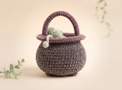 A crocheted miniature pot or cauldron-shaped basket with a rounded body, short feet, and a curved handle, shown on a soft beige background with decorative greenery.