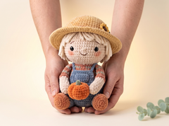 A crocheted amigurumi doll with a straw hat and blue overalls is held in two hands against a warm beige background. The doll holds a small orange pumpkin and has cream-colored yarn hair and face, with blue, orange, and pink yarn details.