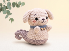 A crocheted plush dog toy with a cream-colored body, floppy ears, a blue collar, and a gray tail, posed against a soft beige background with a eucalyptus leaf branch in the upper left corner.