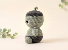 A handmade crocheted amigurumi doll with a light sage green body and a dark charcoal gray hat and lower body, sitting against a warm beige background with soft green eucalyptus leaves in the corners.