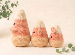 Three cute crocheted candy corn amigurumi toys in different sizes, arranged on a warm beige background with soft green eucalyptus leaves in the corners.