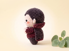 A crocheted doll figure with a dark charcoal knit hat and scarf, cream-colored face and hands, and a burgundy scarf, standing on a warm beige background next to green eucalyptus leaves.