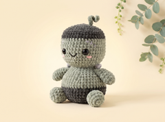 A cute crocheted plush toy shaped like a smiling acorn or nut, sitting on a warm beige background with eucalyptus leaves in the corner. The toy is made of textured yarn in shades of sage green and dark gray, with embroidered facial features.