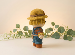 A crocheted scarecrow doll stands on a light beige surface with eucalyptus branches behind it. The doll wears a yellow hat with a brown band, yellow hair, blue overalls, and brown shoes, photographed in a soft studio setting.