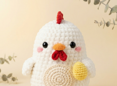 A white crocheted chicken plush toy with a red comb and wattle, pink cheeks, a yellow egg in one wing, and a round beige belly patch, photographed against a soft beige background with blurred eucalyptus branches.