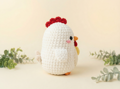 A white crocheted chick toy with a red comb and wattle, standing on a light beige surface with soft-focus green eucalyptus leaves in the background.