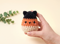 A hand holding a small crocheted cat character toy with a black top, orange middle, and cream bottom, against a pale yellow background with a green eucalyptus sprig in the corner.