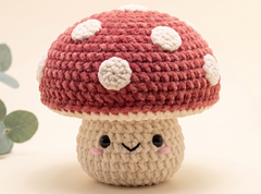 A cute crocheted mushroom plush toy with a dusty red cap, white spots, and a beige stem with embroidered facial features, set against a soft cream background with a blurred green leaf on the left.