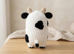 A crocheted plush cow toy with black spots and beige horns, standing on a wooden surface against a soft beige background.