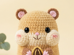 A close-up of a crocheted hamster plush toy with a beige body, cream-colored cheeks, brown ears, pink inner ears and nose, and black bead eyes, set against a soft cream background.