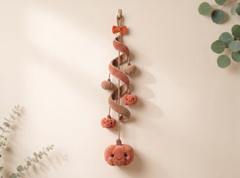 A knitted autumn hanging decoration with a spiral brown ribbon, small orange pumpkins, and a bow at the top, displayed against a warm beige wall with soft eucalyptus leaves in the corners.