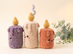 Three cute crocheted candle-shaped plush toys in purple, cream, and terracotta orange, arranged on a warm beige background with a sprig of eucalyptus.