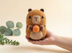 A crocheted beaver toy held in a person's hand against a warm beige background, with decorative green leaves in the corner.