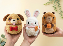 Three cute crocheted plush toys are held in hands against a soft beige background with green leaves. The toys are shaped like a tan dog with a red bow tie, a white bunny holding an orange carrot, and a brown beaver holding an orange acorn.