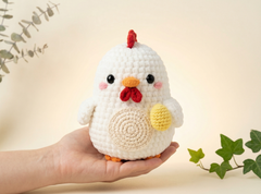 A crocheted white chicken amigurumi toy with a red comb and wattle, orange beak and feet, and a yellow wing, held in a person's hand against a warm cream background with green leaves.