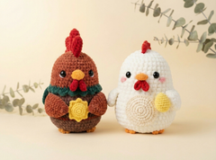 Two crocheted chicken amigurumi toys standing side by side on a light beige surface. One is brown with a red comb, yellow beak and egg detail, and a green scarf.