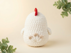 A crocheted white chick toy with a small red comb and beige feet, photographed on a warm cream background with soft green leaves in the corners.