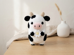 A crocheted cow figurine with black and white patches, a pink snout, and small tan horns, standing on a wooden surface with a soft neutral background and a small white vase behind it.