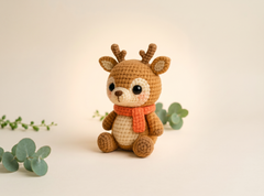 A crocheted plush deer toy with brown antlers and legs, a cream-colored belly and face, and an orange scarf, sitting on a warm beige background with decorative eucalyptus leaves.
