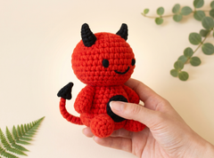 A small crocheted devil plush toy in bright red with black horns, tail tip, and belly patch, held in a person's hand against a warm cream background with green leafy stems.