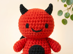 A red crocheted amigurumi doll with black horns, black eyes, a black stitched smile, and a black belly patch, photographed against a soft cream background with a small green plant visible in the upper right.